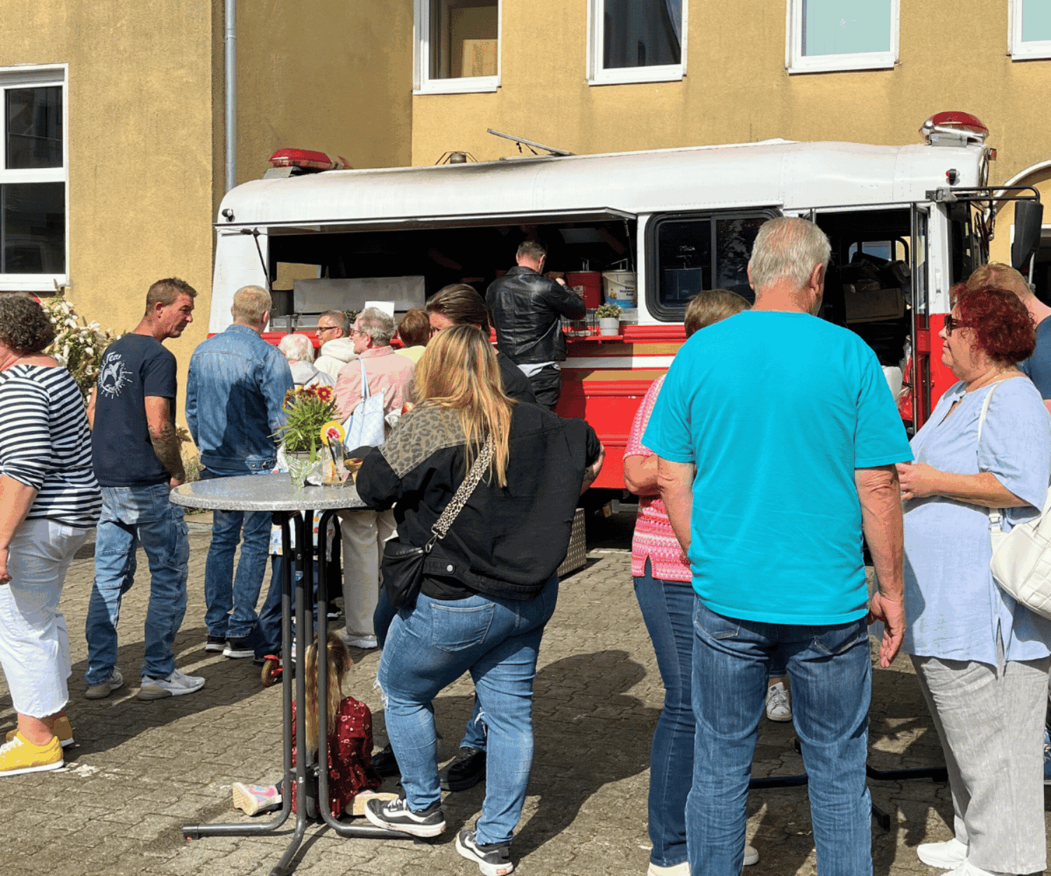 Das Bild zeigt das Sommerfest der Familien- und Krankenpflege Bochum. Eine Gruppe von Menschen stehen an Stehtischen an einem Foddtruck. Die Sonne scheint.