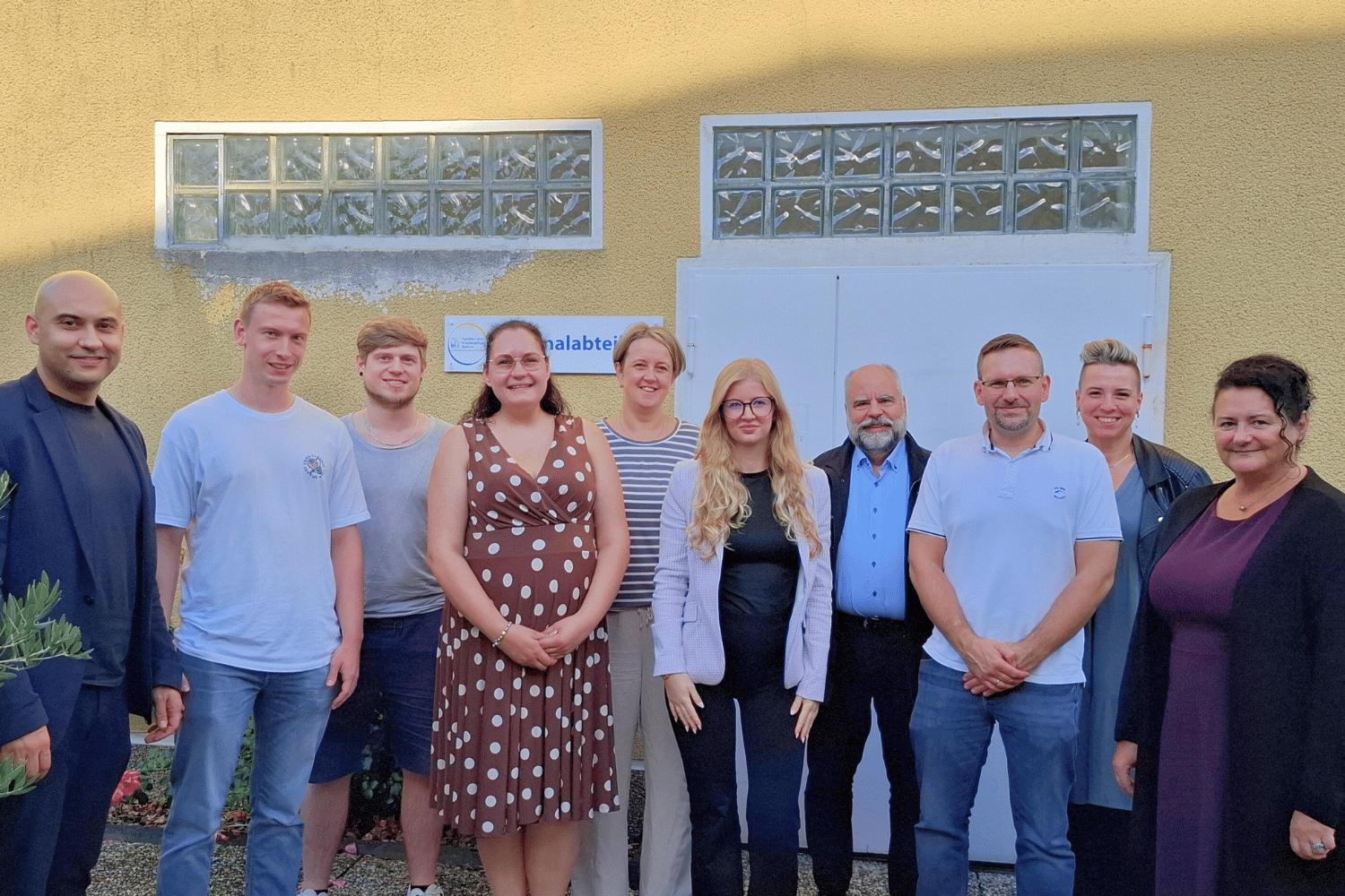 Das Bild zeigt eine Gruppe von Menschen, insgesamt 10 Personen.Davon sind 5 Frauen und 5 Männer.Die Personen stehen vor einer gelb verputzten Hauswand und lächeln freundlich in die Kamera.