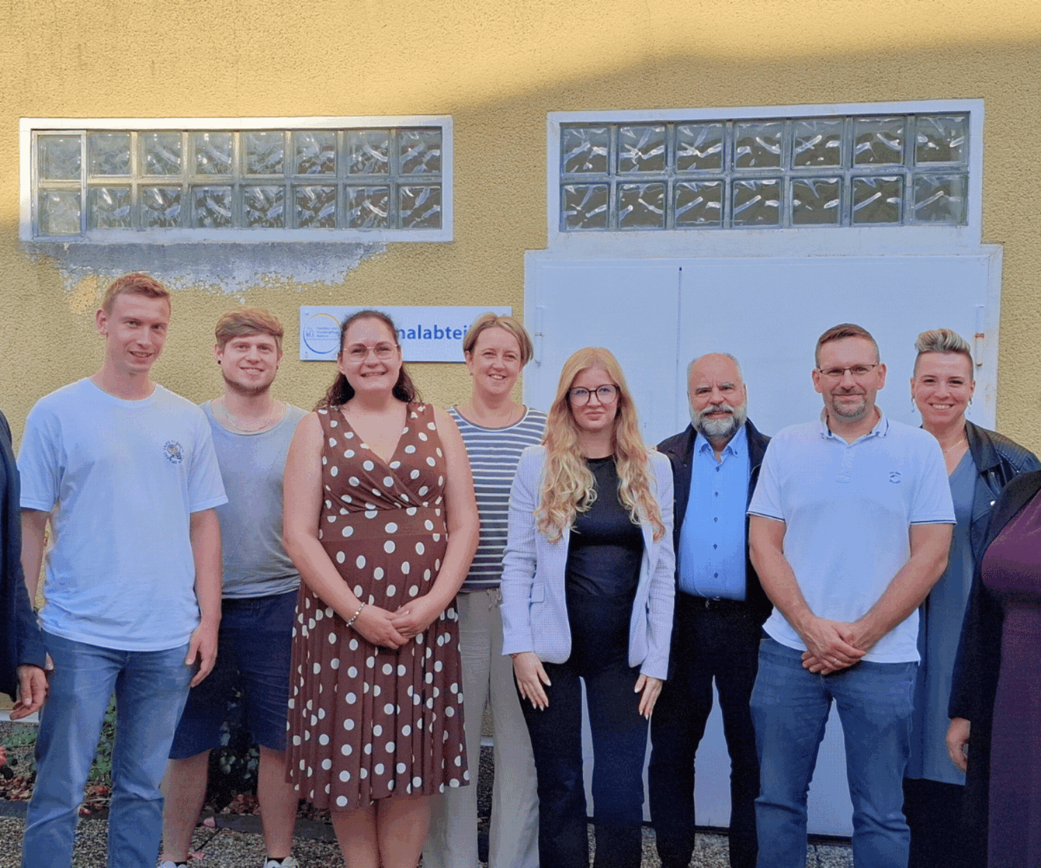 Das Bild zeigt eine Gruppe von Menschen, insgesamt 10 Personen.Davon sind 5 Frauen und 5 Männer.Die Personen stehen vor einer gelb verputzten Hauswand und lächeln freundlich in die Kamera.