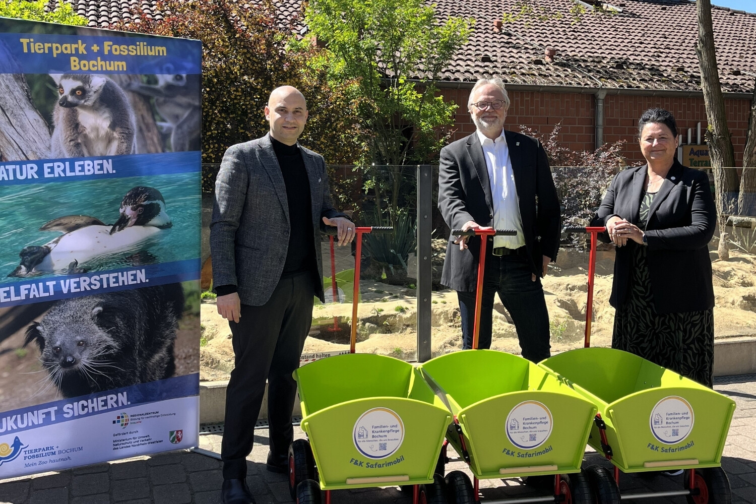 3 Personen stehen mit grünen Bollerwagen bei Sonnenschein im Bild.
