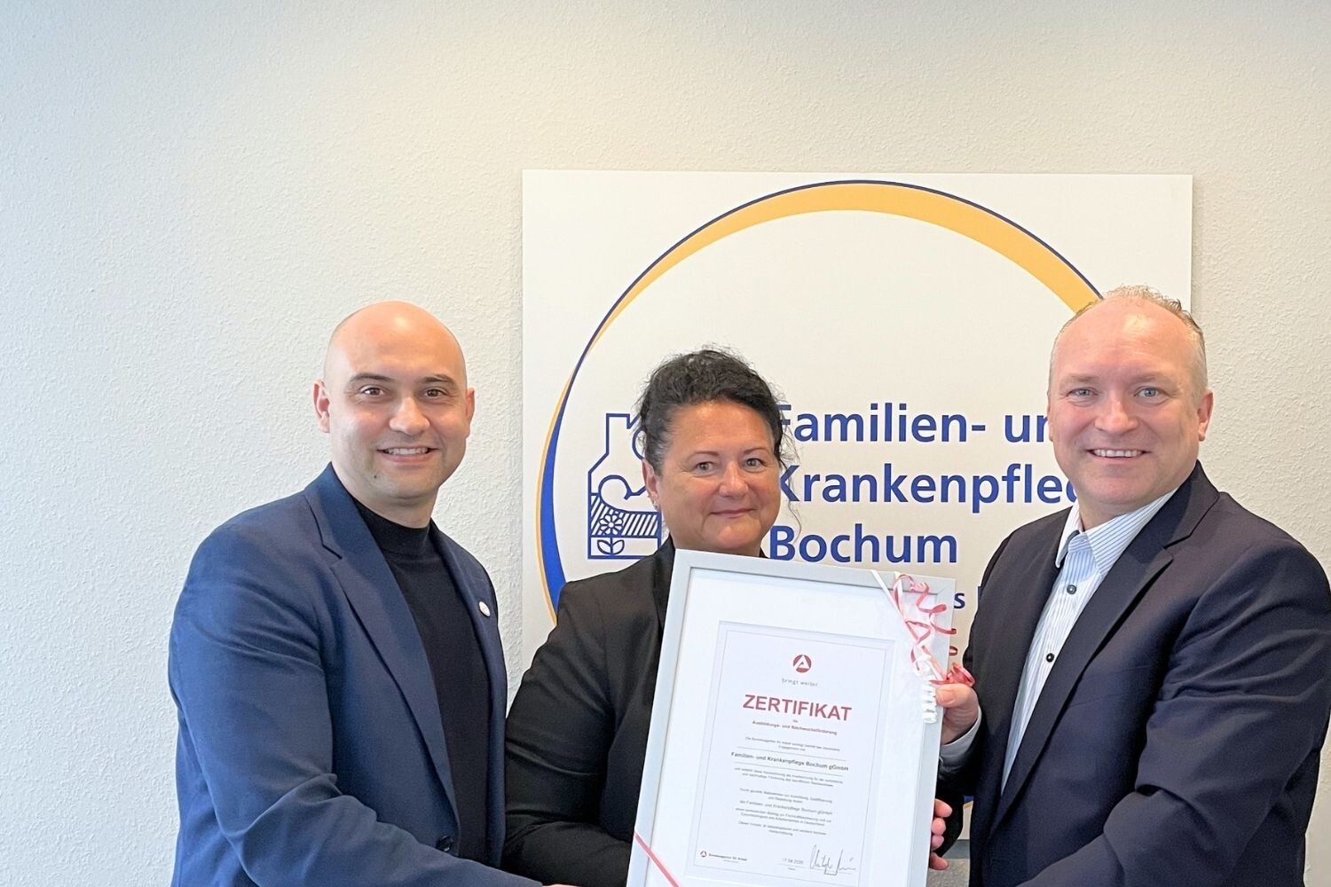 3 Personen sind auf dem Bild. In der Mitte steht eine Frau während rechts und links jeweils ein Mann stehen. Das Bild zeigt die feierliche Übergabe einer Urkunde. Im Hintergrund sieht man das Logo der Familien- und Krankenpflege Bochum.