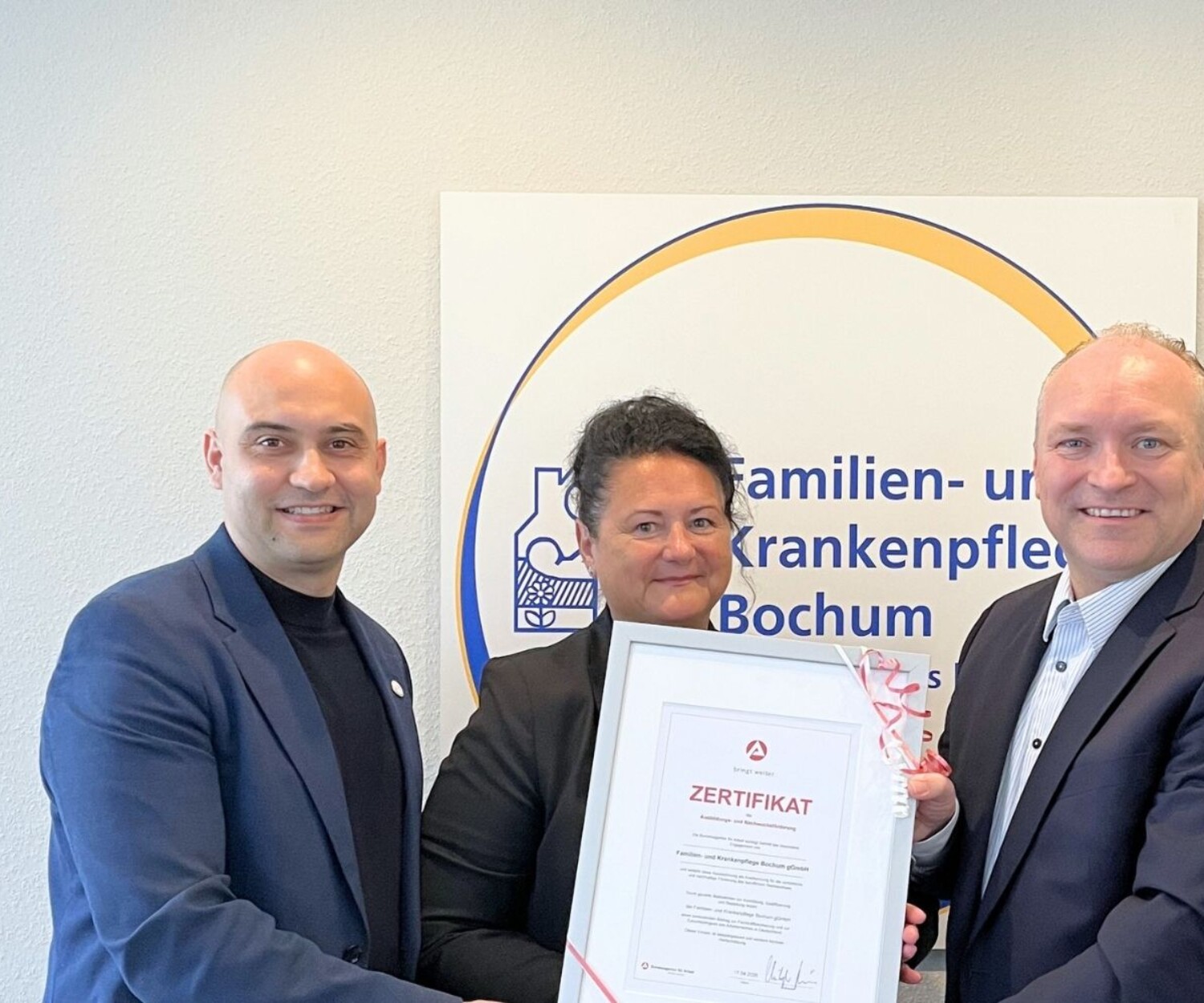 3 Personen sind auf dem Bild. In der Mitte steht eine Frau während rechts und links jeweils ein Mann stehen. Das Bild zeigt die feierliche Übergabe einer Urkunde. Im Hintergrund sieht man das Logo der Familien- und Krankenpflege Bochum.